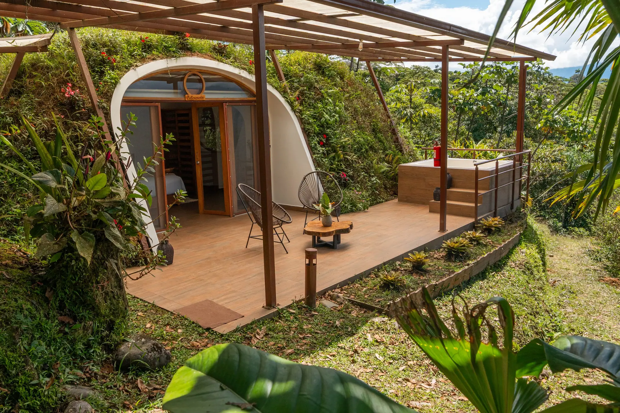 Un domo en la selva con frente de vidrio en arco bajo una pérgola de madera, dos sillas en su terraza de madera y una ladera de plantas alrededor