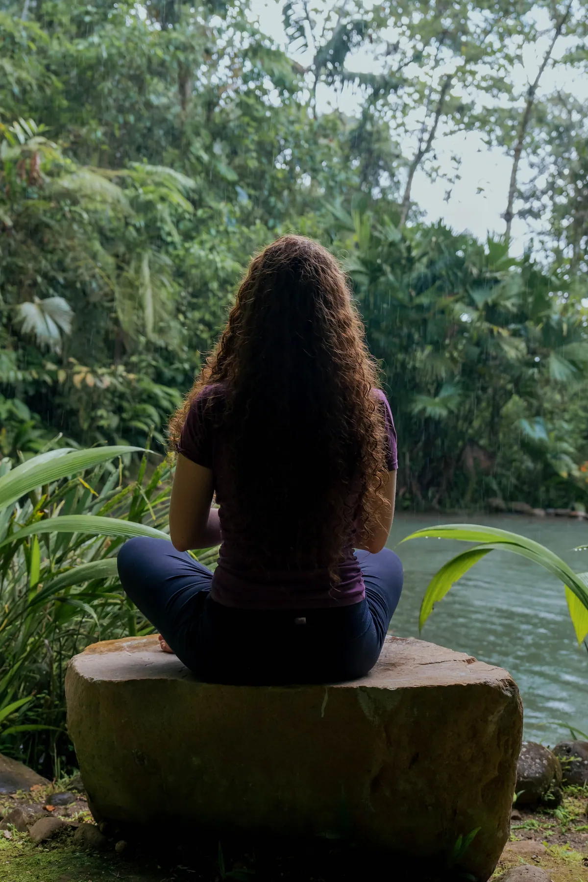 Una mujer sentada con las piernas cruzadas sobre una piedra plana encima de un río verde, de espaldas a la cámara, con lluvia cayendo entre las palmeras