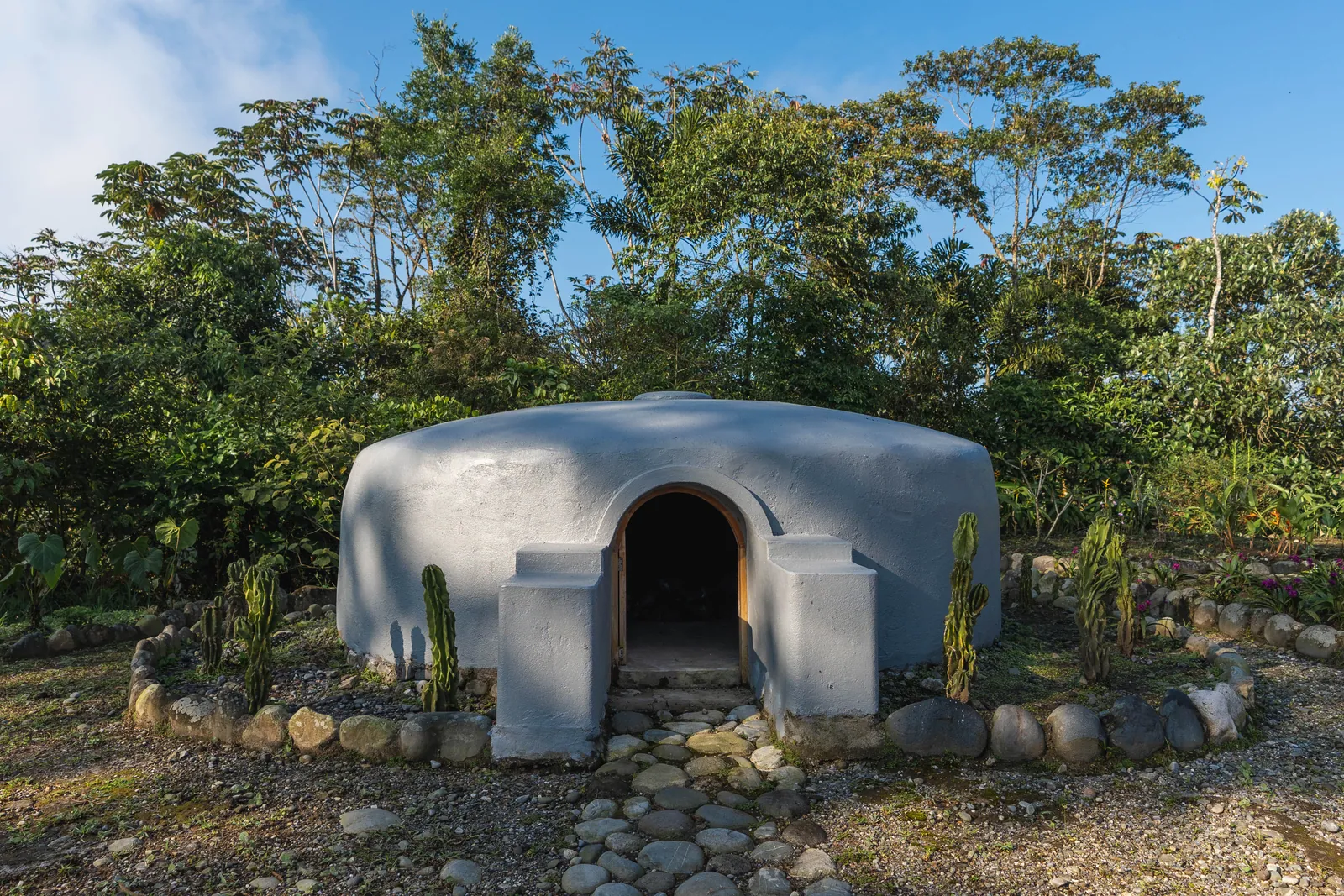 Un temazcal bajo y abovedado, de piedra revocada clara y puerta en arco, rodeado de piedras de río y cactus bajo el dosel del bosque