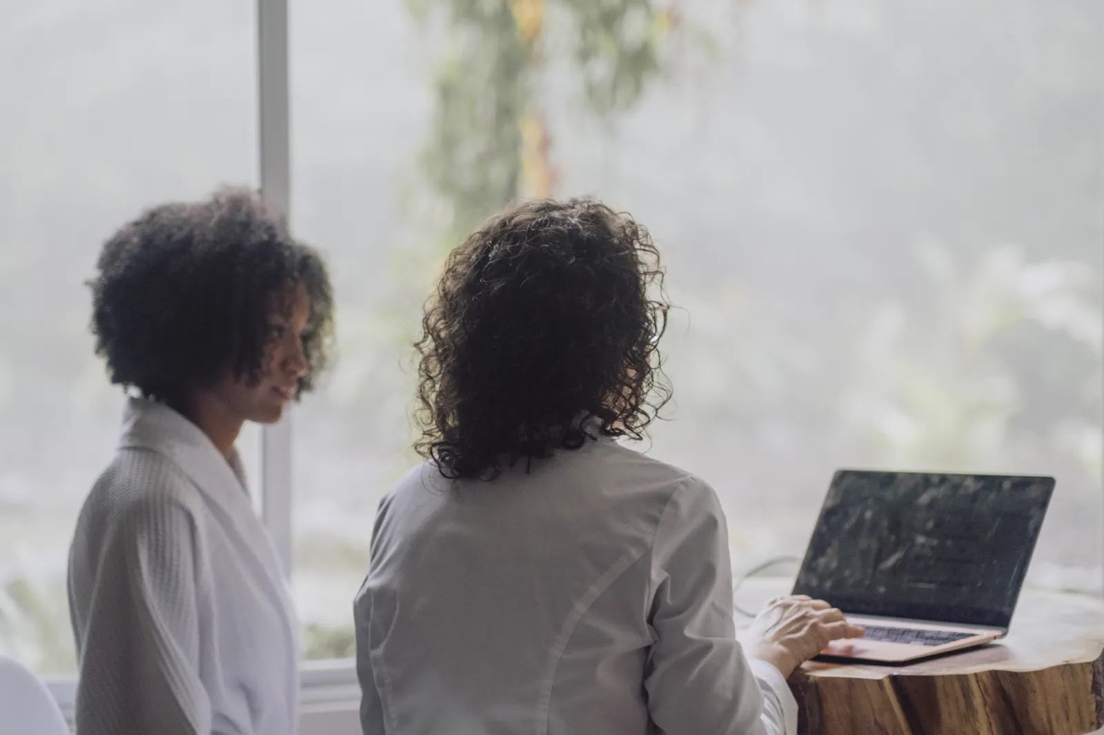 Una huésped en bata y la médica del lodge en una mesa de madera, sus lecturas abiertas entre ellas, el bosque nublado en la ventana del fondo