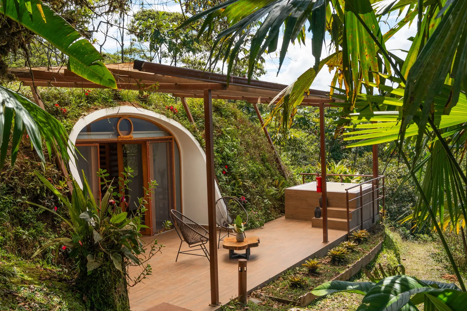 Un domo en la selva con frente de vidrio en arco, dos sillas y una tina de madera en su terraza techada, rodeado de palmeras y helechos