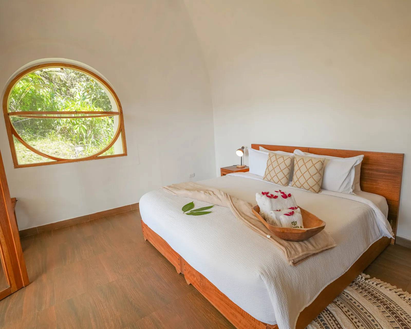 Dentro de un domo: una cama con toallas dobladas y hojas sobre el cubrecama, y una ventana redonda de madera hacia el jardín